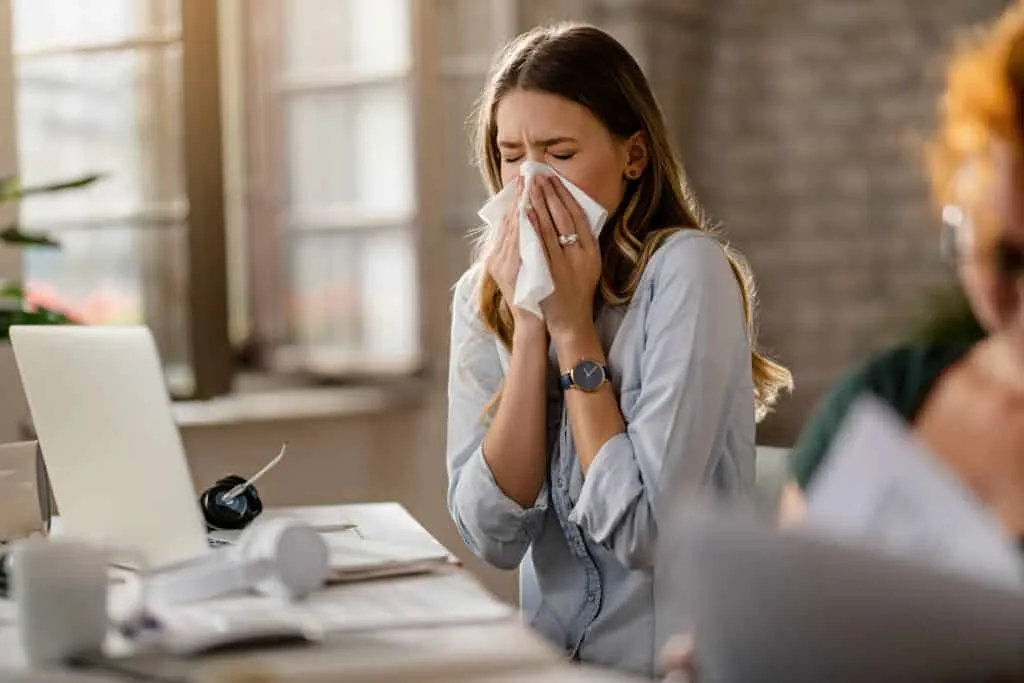 young sick businesswoman sneezing tissue while working office scaled